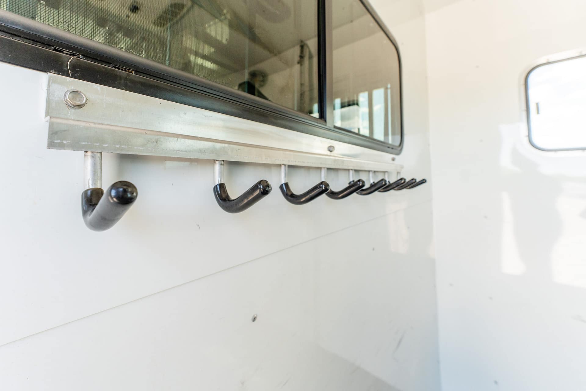 Wall-mounted metal rail with black curved hooks on a white wall, corner of a window visible in the background.