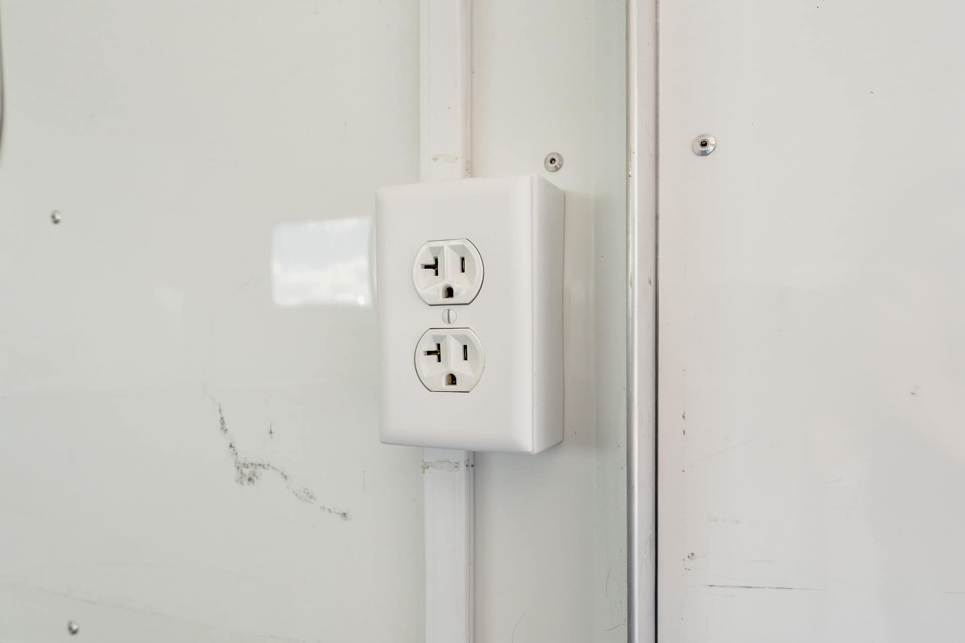White duplex wall outlet with two three‑prong receptacles mounted on a light wall near a vertical metal rail portion.