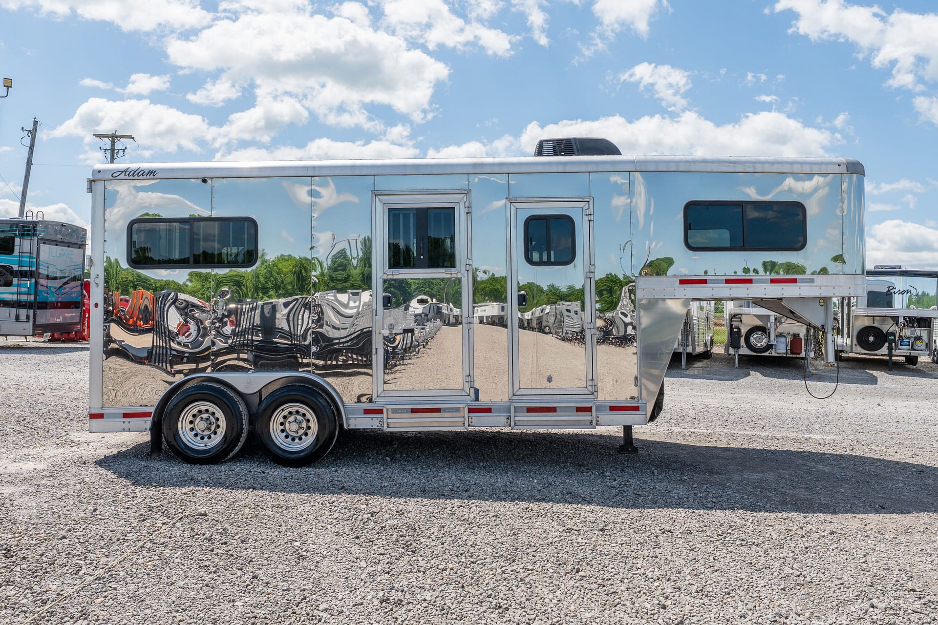 Shiny chrome trailer with mirrored sides labeled 'Adam', reflecting surrounding vehicles in a gravel lot under a blue sky with clouds.