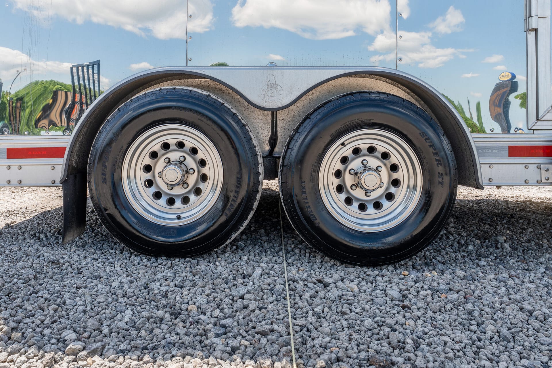 Close-up of twin trailer wheels on gravel under a shiny metal trailer, with a thin rope on the ground.