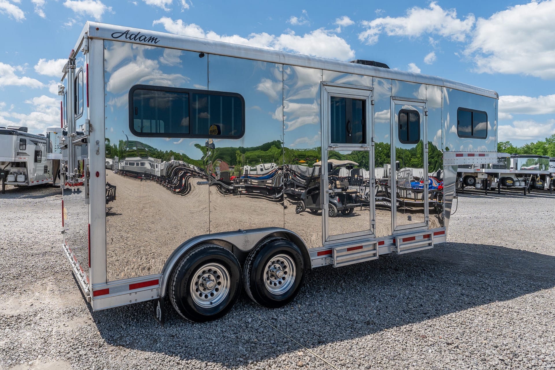 Shiny aluminum trailer with reflection of other trailers and blue sky, parked on a gravel lot.