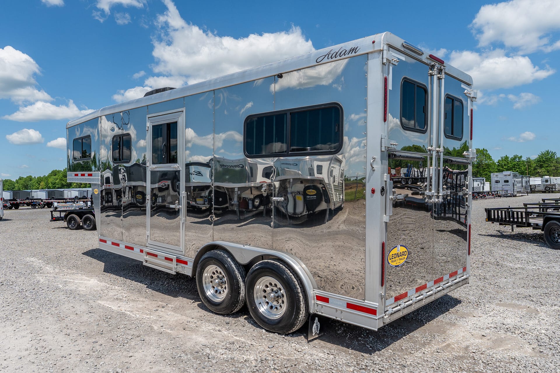 Polished aluminum cargo trailer with windows parked on a gravel lot under a blue sky, with other trailers in the background