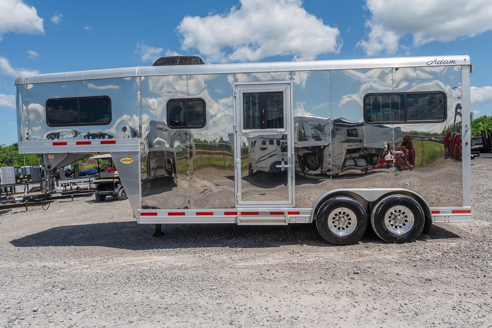 Shiny aluminum travel trailer with two side windows and a central door, parked on a gravel lot under a blue sky with clouds.