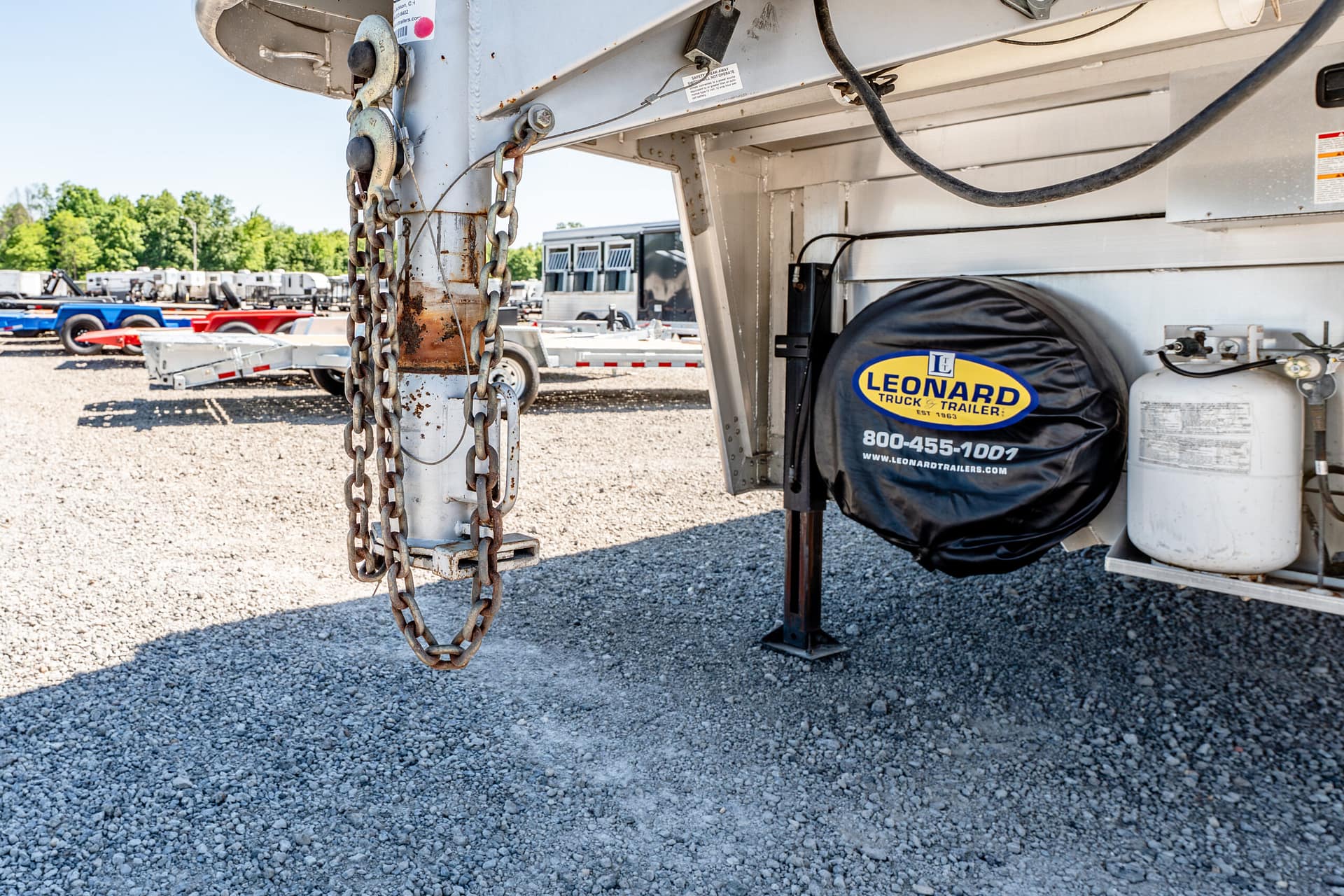 Close-up of a trailer hitch with rusted chains hanging, a Leonard spare tire cover, and a propane tank in a gravel lot.