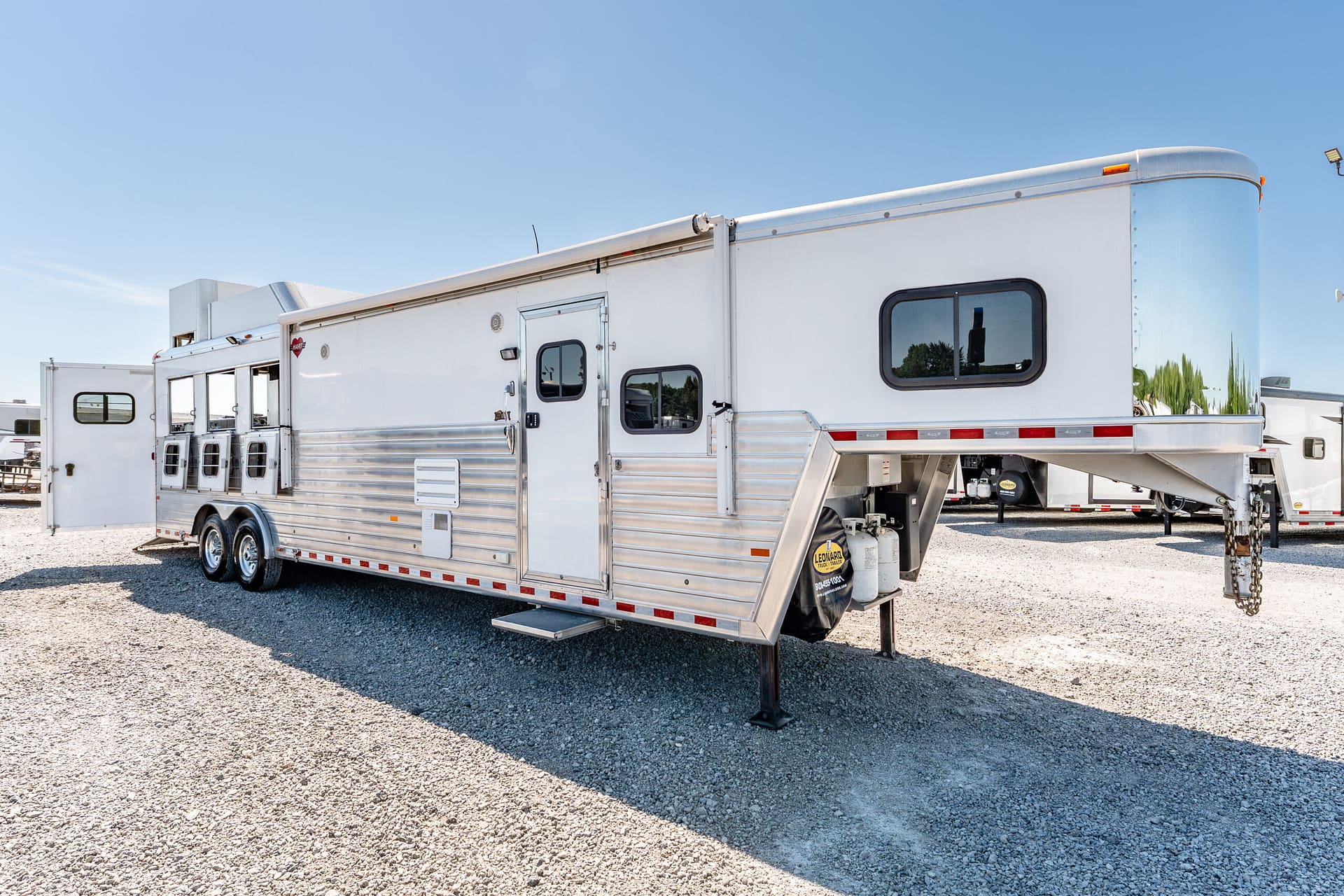 Shiny silver aluminum horse trailer on a gravel lot under a clear blue sky, with windows and a ramp visible on the side.