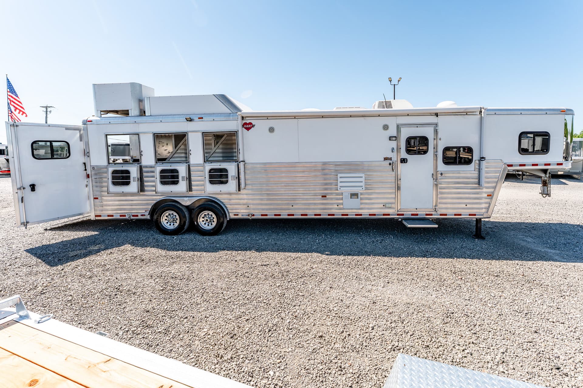 Long white horse trailer with multiple windows and dual axles parked on a gravel lot under a clear blue sky; an American flag is visible in the background.