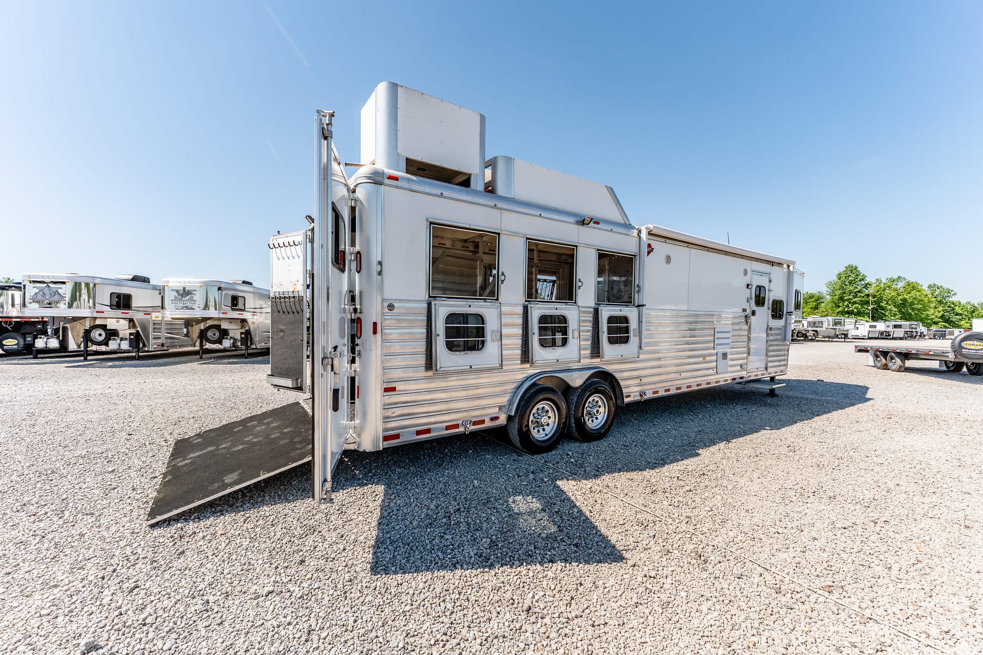 Silver horse trailer with ramp down, parked on a gravel lot under a clear blue sky.