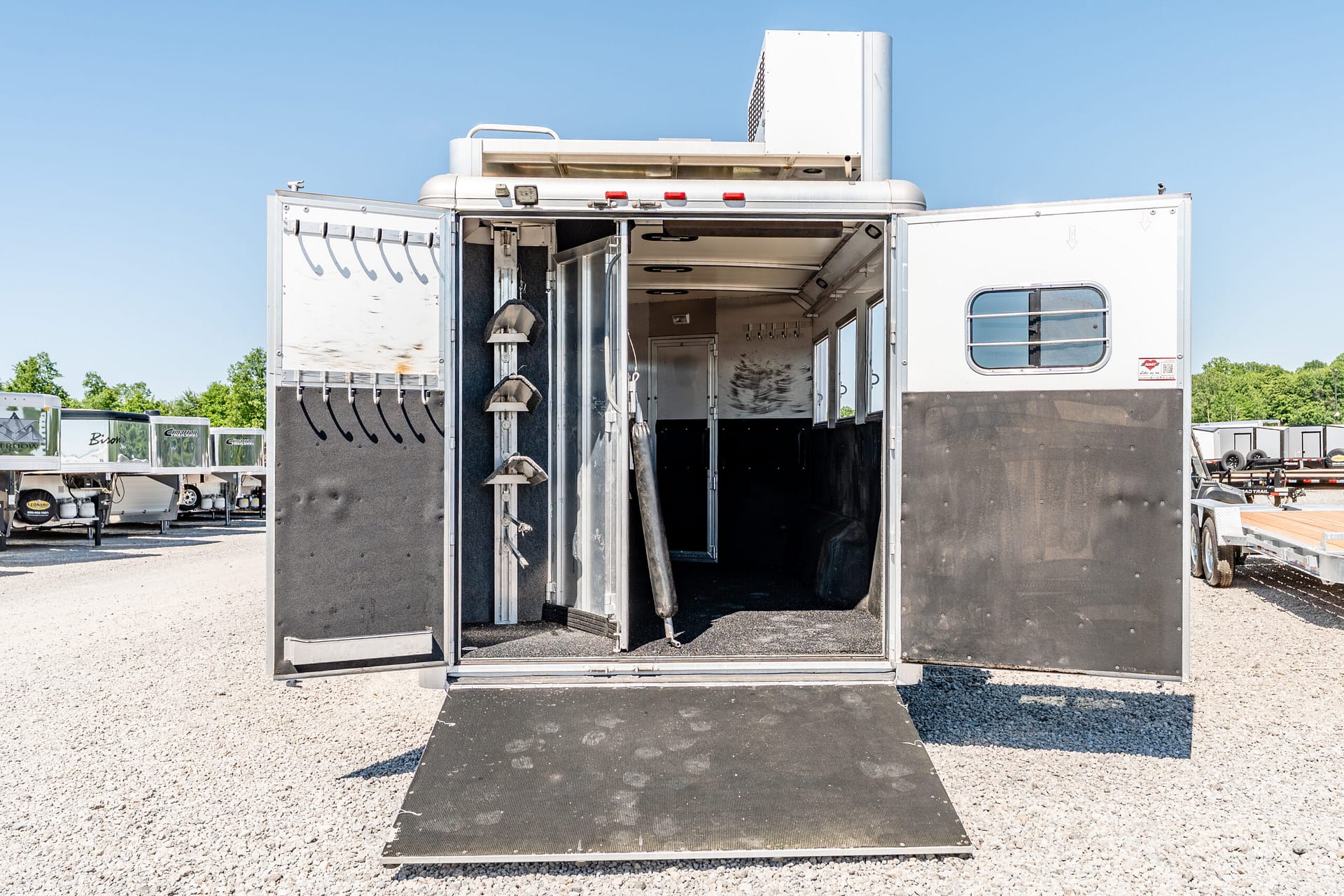 Open horse trailer with lowered ramp, showing tack racks on the left and empty stalls inside, parked on a gravel lot with other trailers nearby under a clear blue sky.