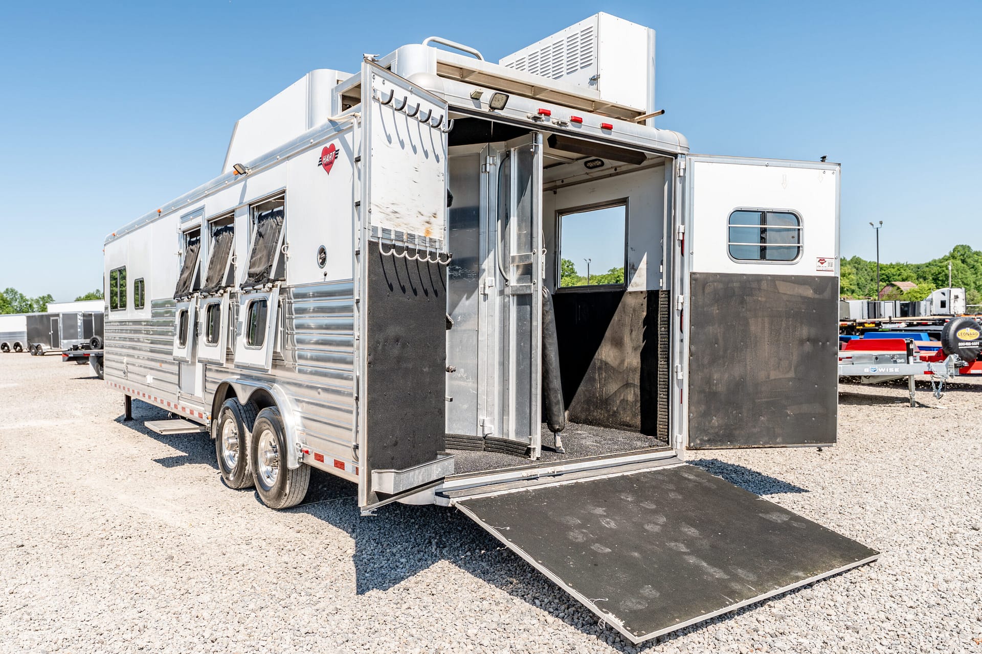 Silver horse trailer with a lowered rear ramp, exposing the interior stalls and loading area on a gravel lot under a clear blue sky.