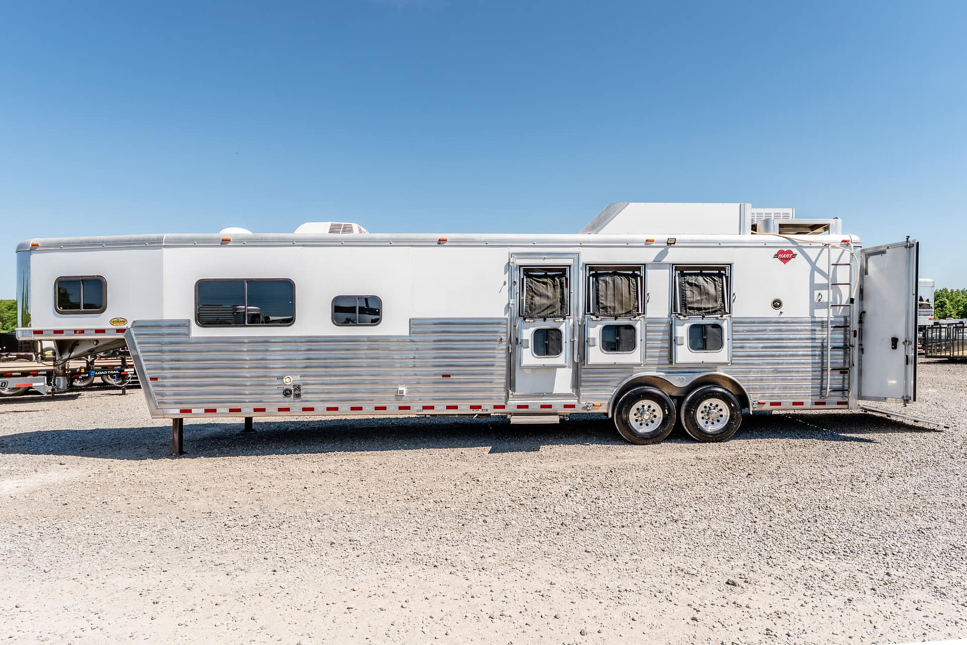Silver travel trailer parked on a gravel lot under a clear blue sky, side view with multiple windows and doors