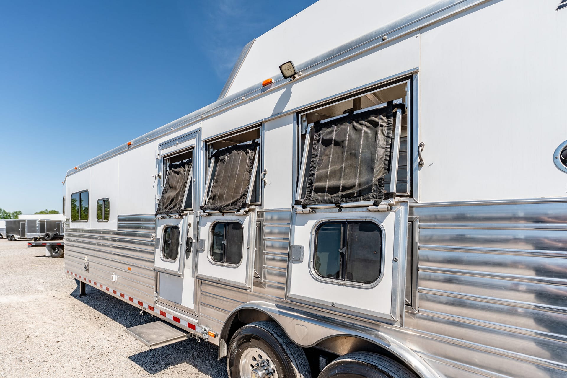 Silver aluminum horse trailer with open windows and black curtains, parked on a gravel lot under a blue sky.