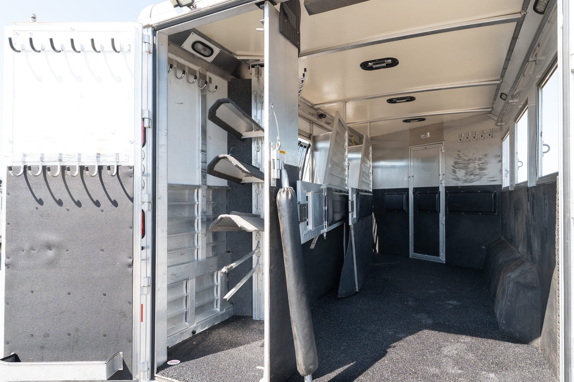 Interior of a metallic horse trailer with a central divider, stair steps on the left, and coat hooks along the wall inside the doorway