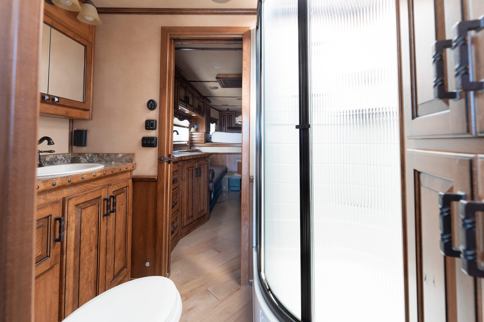 Interior of an RV bathroom with wood cabinets, a sink, and a curved glass shower door in the background on the right side.