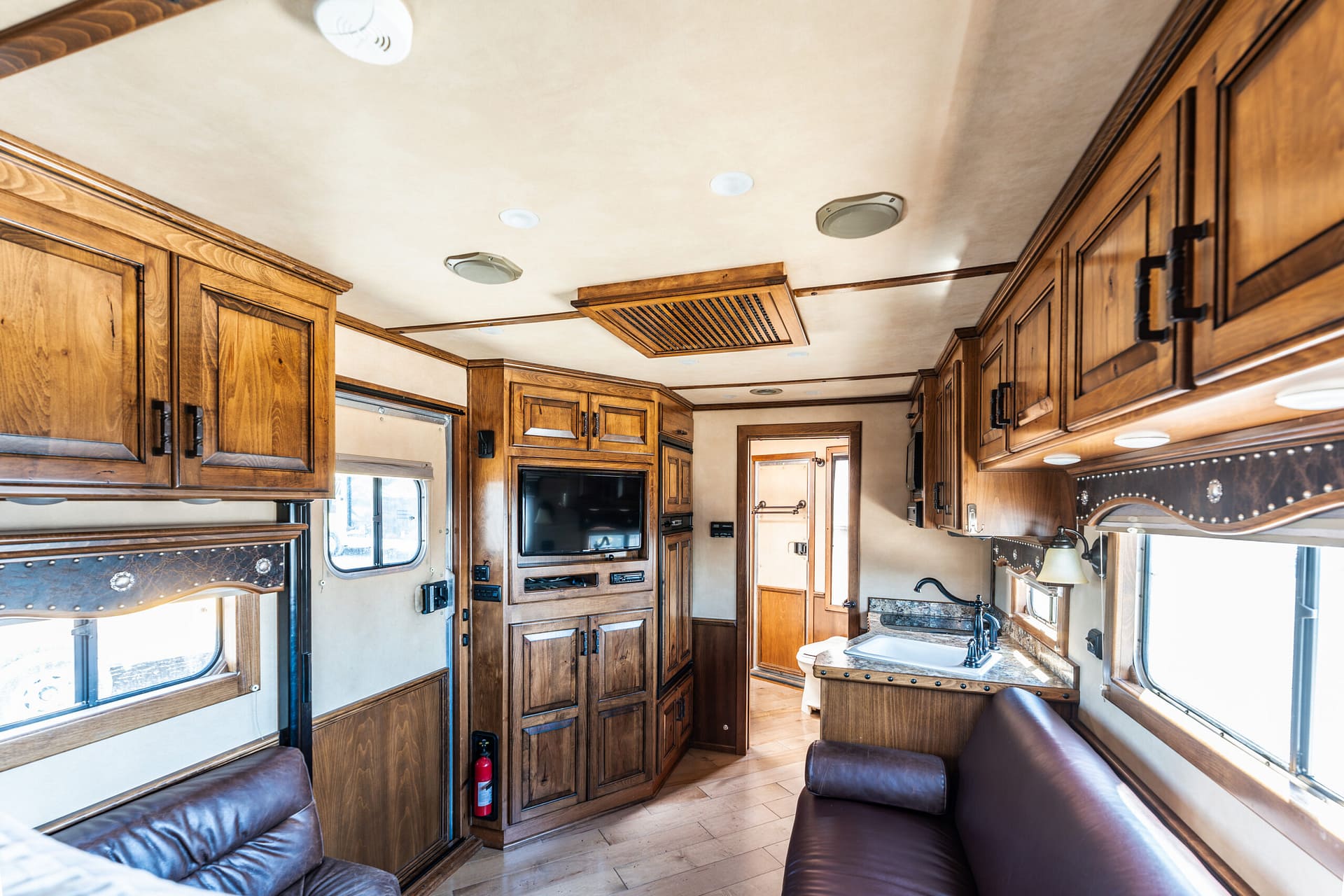 Interior of a luxury RV: brown wood cabinets flank the room with leather sofas and a small kitchen area visible.