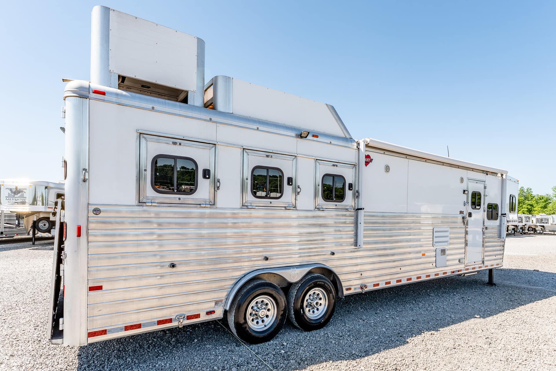 Side view of a silver aluminum horse trailer with multiple windows parked on a gravel lot under a clear blue sky, showing its doors and wheels.