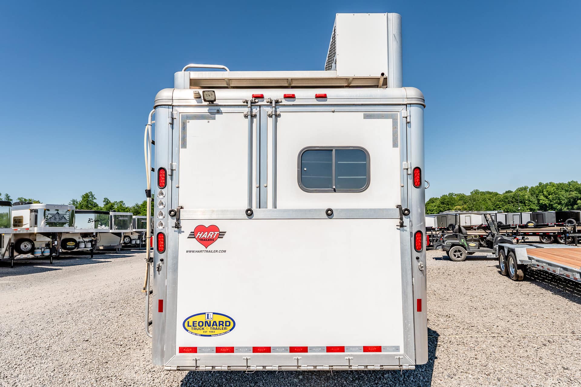 Rear view of a silver Hart trailer in a gravel lot, with a blue sky and other trailers in the background, and Leonard sticker near the bottom left.