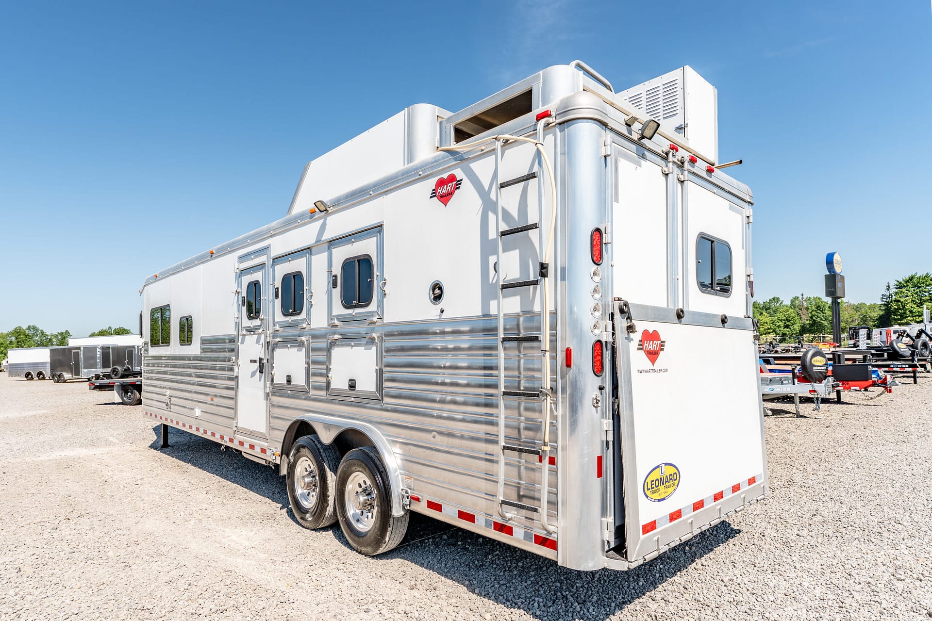 Large silver Hart trailer parked on a gravel lot under a clear blue sky, with other trailers in the background.