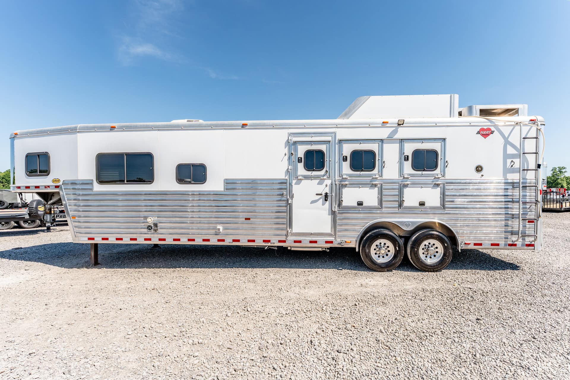 Large white aluminum horse trailer with multiple windows and compartments parked on gravel under a clear blue sky, showing tandem wheels and external storage doors.