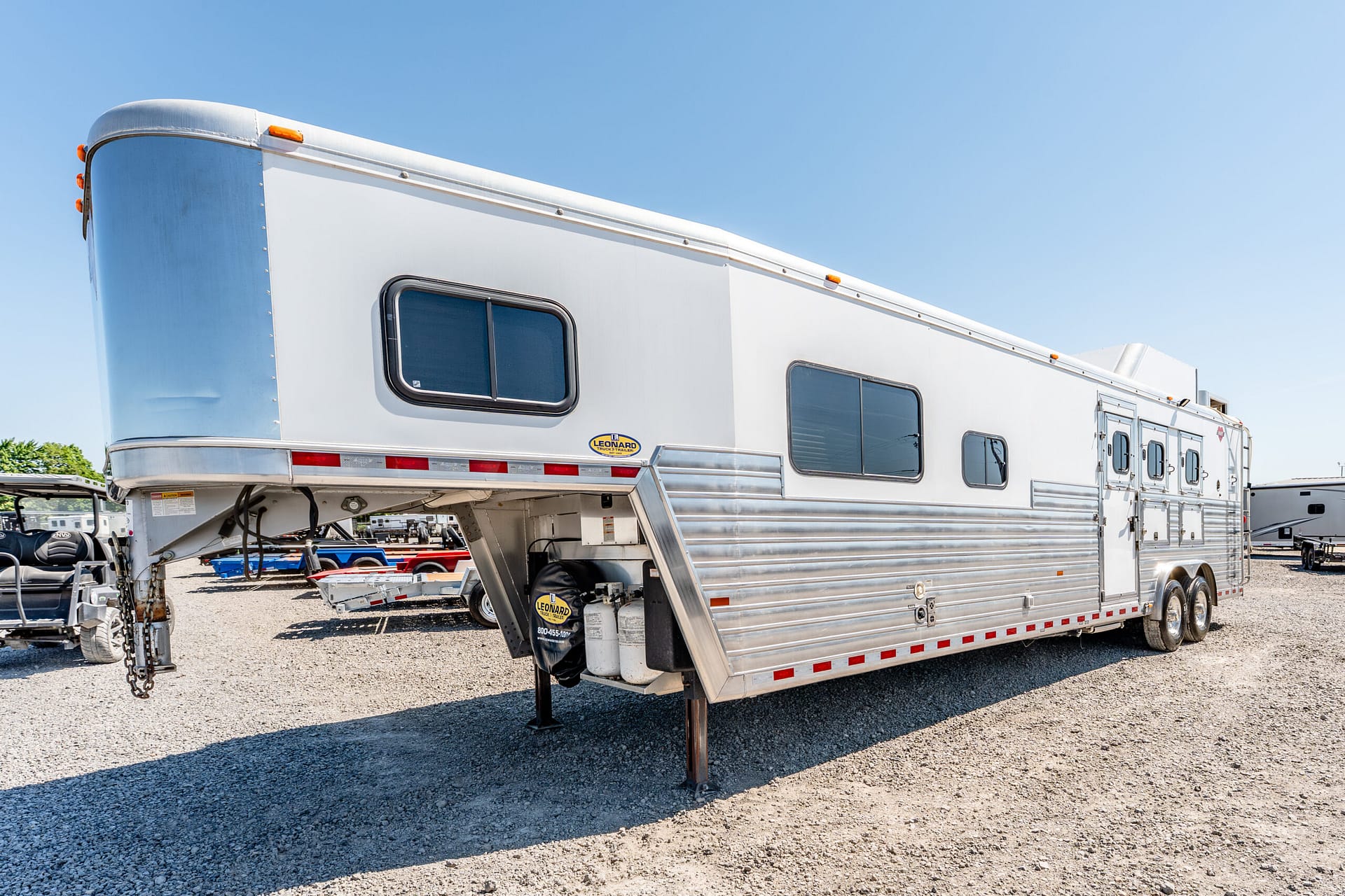 Large silver travel trailer with blue-tinted windows parked on gravel under a clear blue sky, stabilizer legs down.