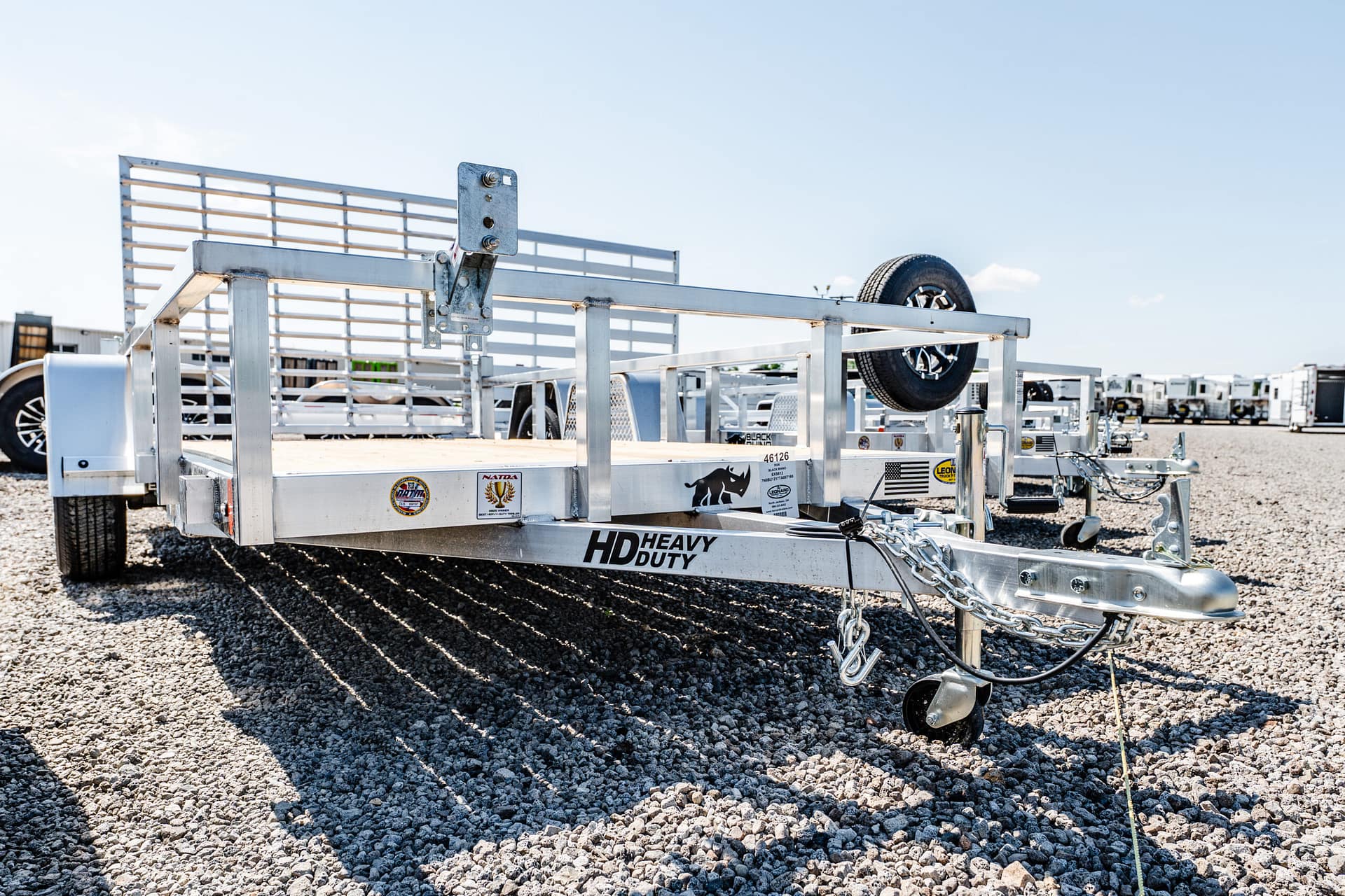 Silver metal utility trailer on a gravel lot with a mounted spare tire and other trailers in the background.