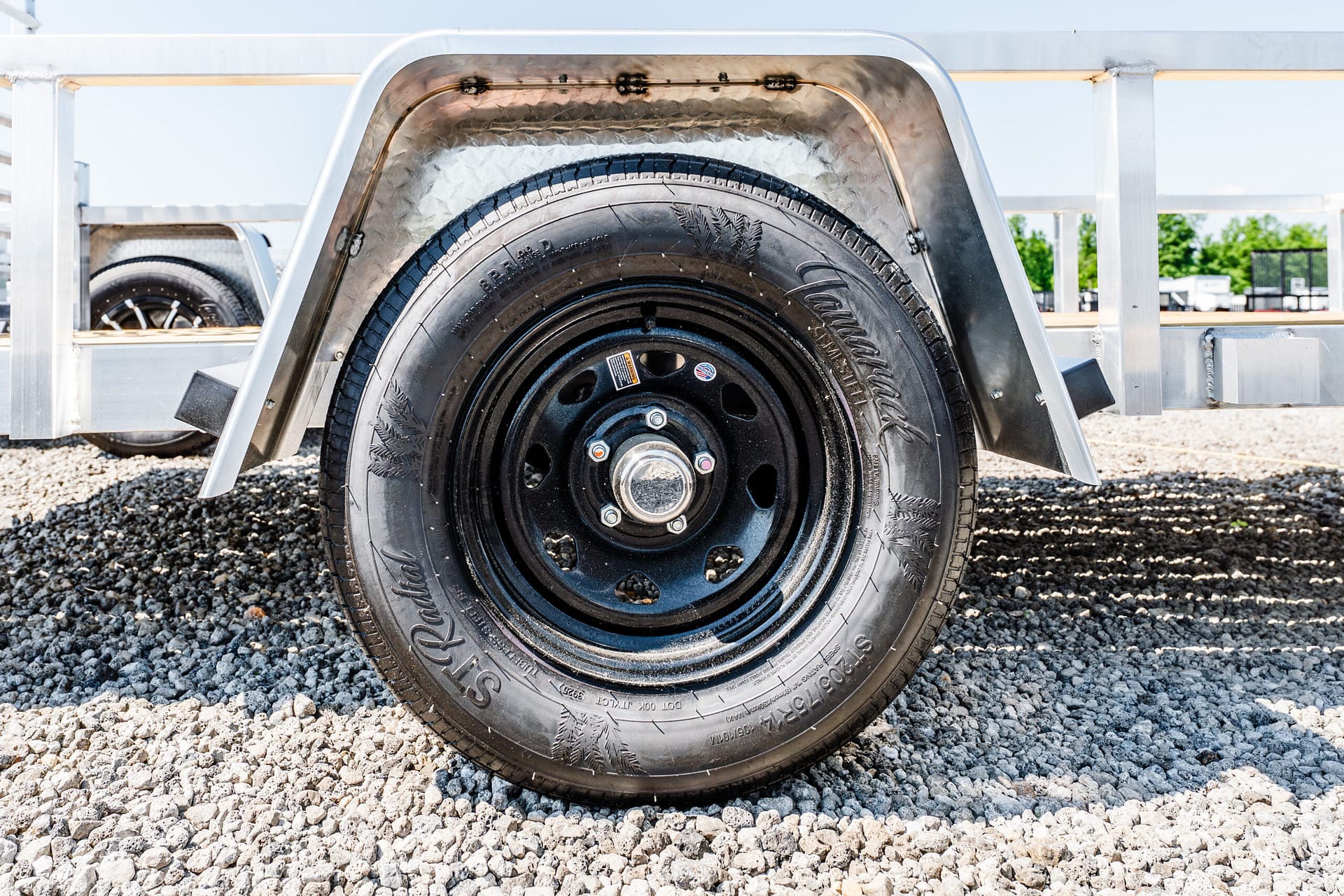 Close-up of a black steel trailer wheel and tire under a curved metal fender on gravel ground.