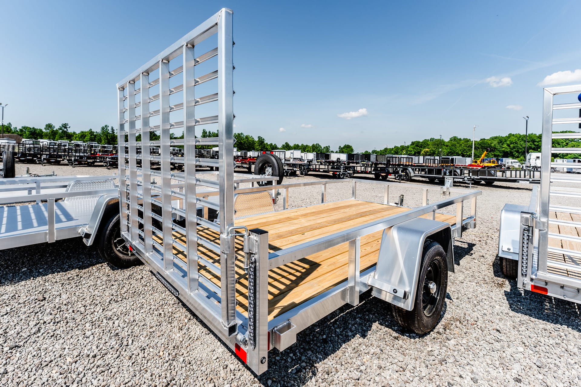 Silver utility trailer with a wooden deck and metal frame, parked in a lot of trailers under a clear blue sky.