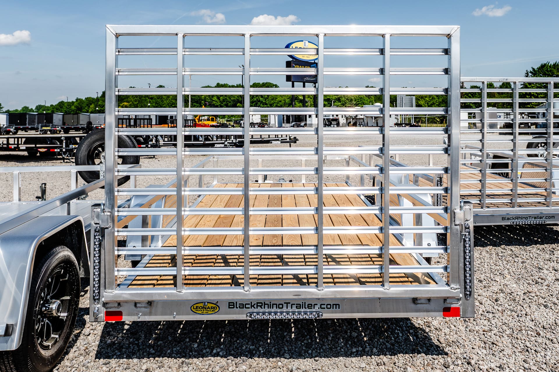 Rear view of a silver utility trailer with a wooden deck and metal slats, in a dealership lot; branding reads BlackRhinoTrailer.com.