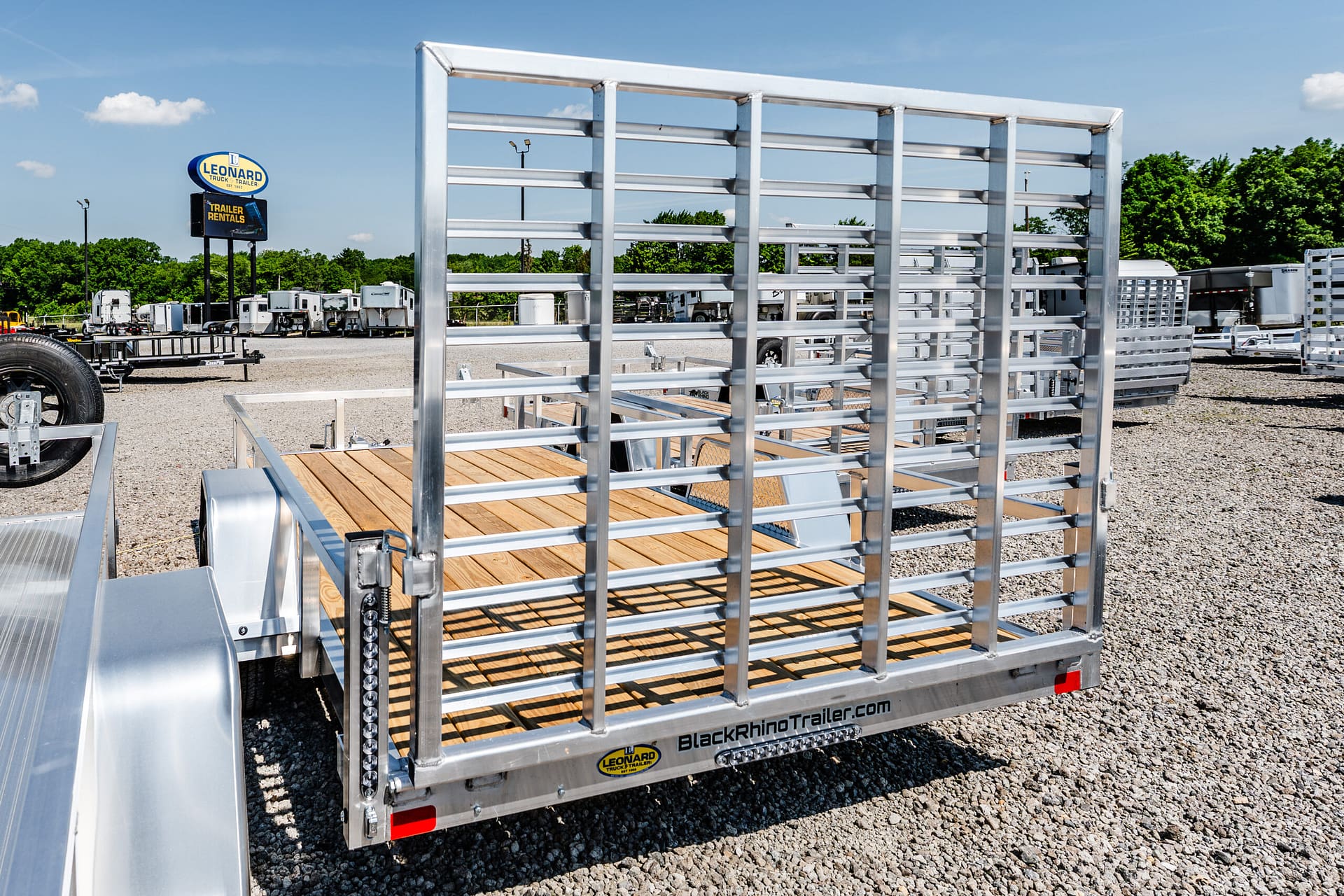 Rear view of a silver metal-frame flatbed trailer with a wooden floor, at a trailer lot with Leonard sign in the background; blue sky.