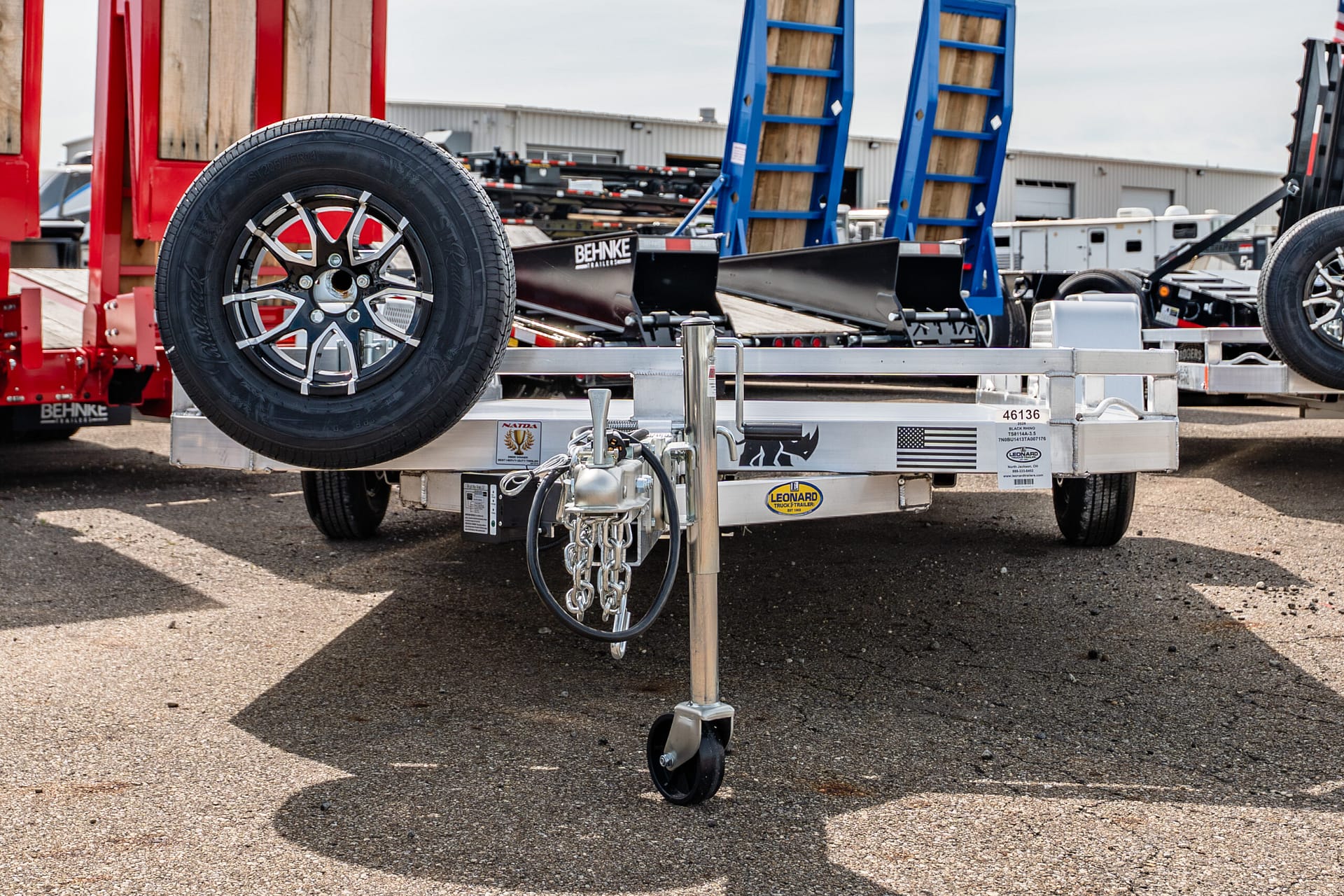 Close-up of a trailer's spare tire mounted on a chrome wheel, with hitch and jack in the foreground at a trailer yard.