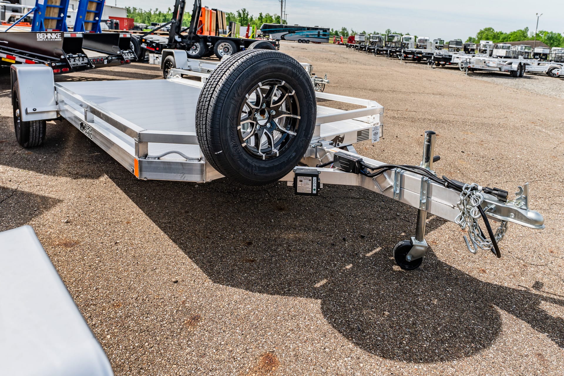 Aluminum flatbed trailer with a spare wheel mounted at the front, parked in a gravel lot among many trailers in the background.