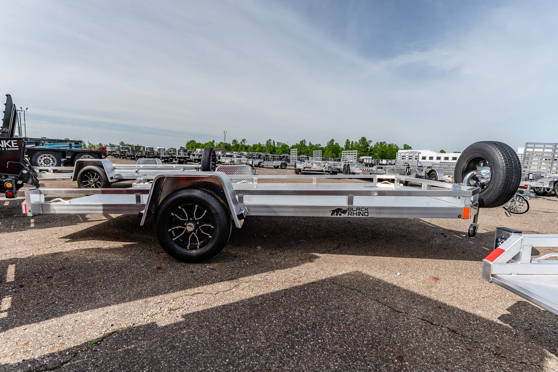 Row of aluminum utility trailers with spare tires parked in a gravel trailer yard under a clear blue sky.