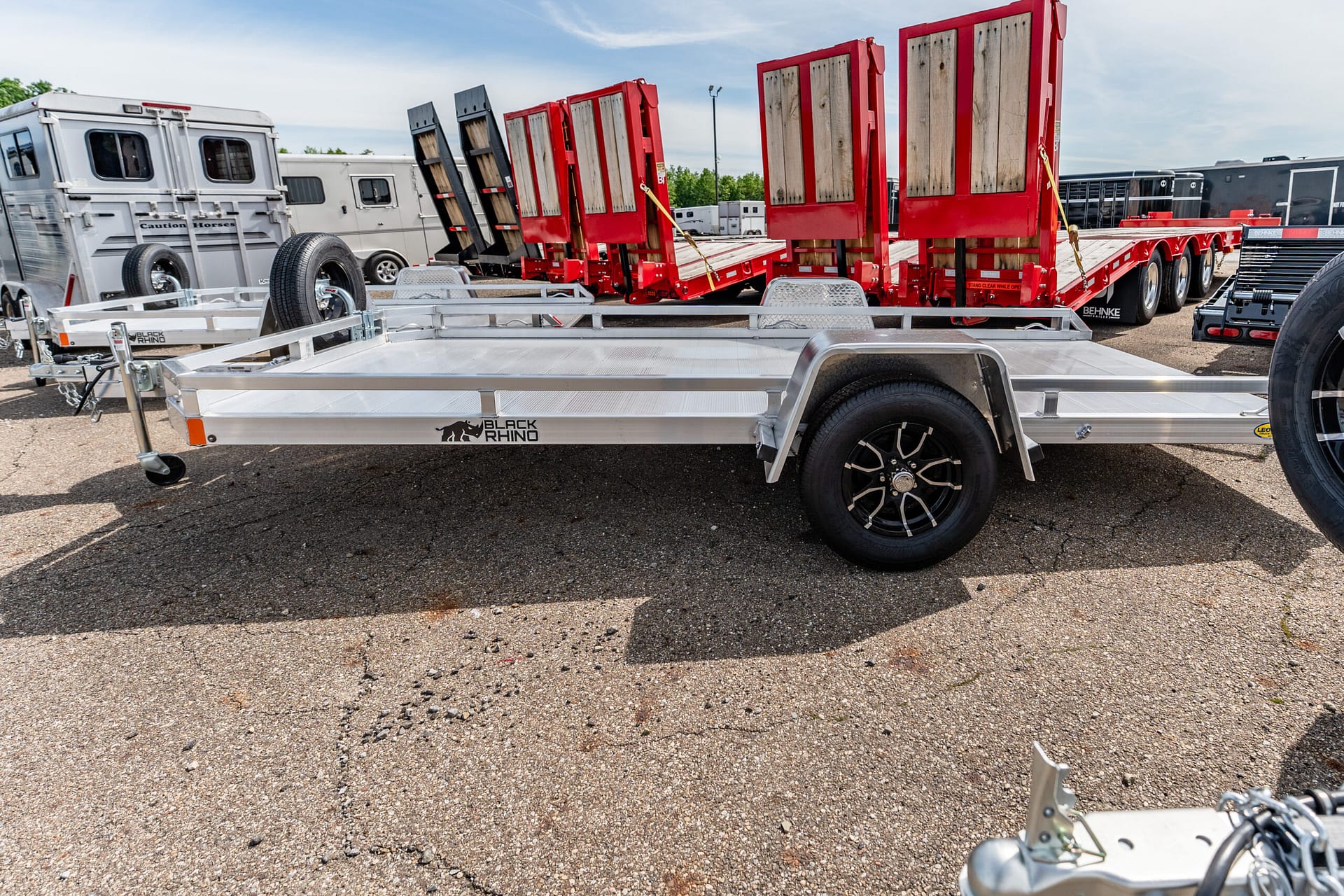 Aluminum flatbed trailer with side rails and tall wooden ramps standing upright in a trailer lot, with other trailers in the background.
