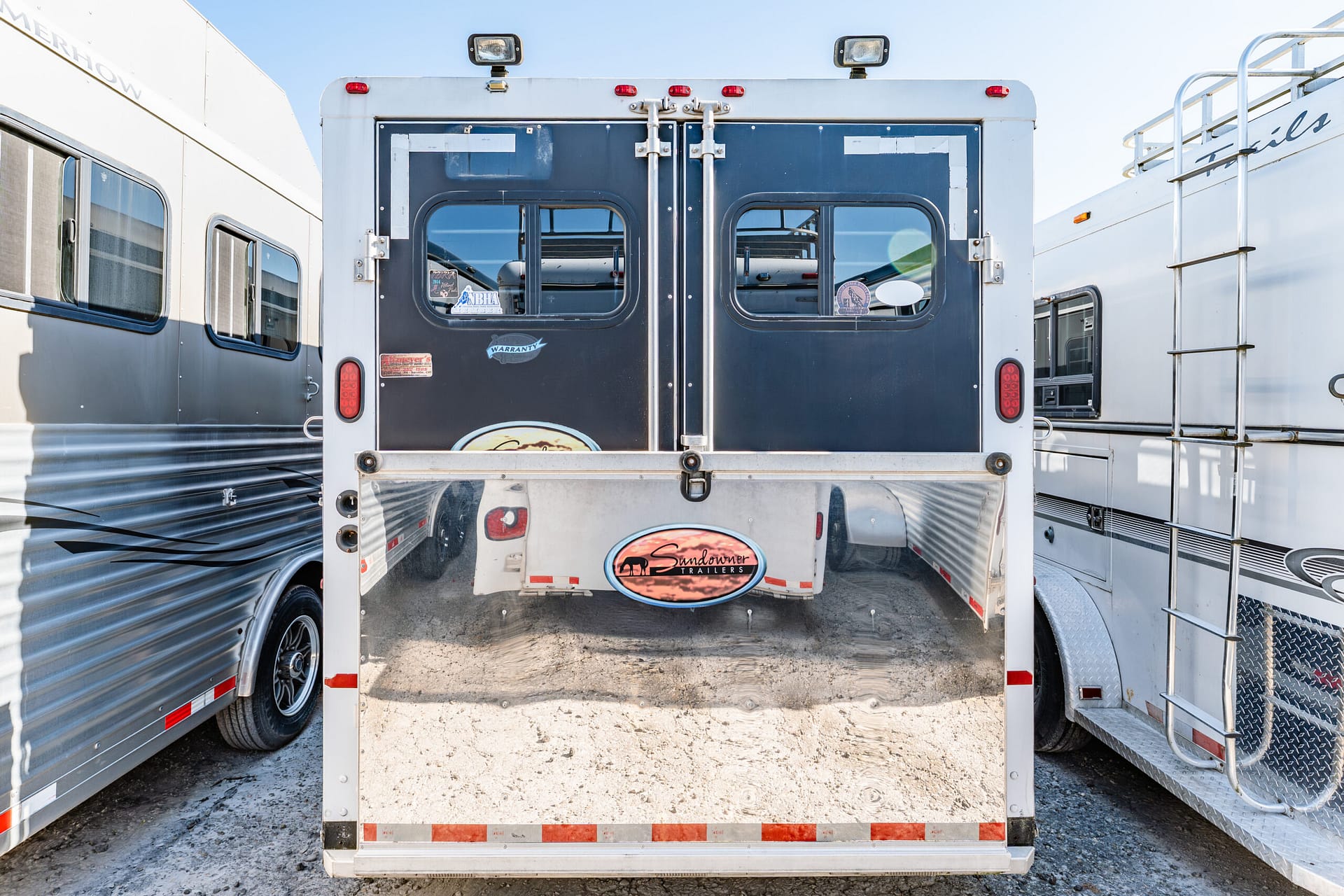 Rear view of a dark blue Sundowner trailer with double doors, windows, and bumper, between silver trailers under a clear sky.