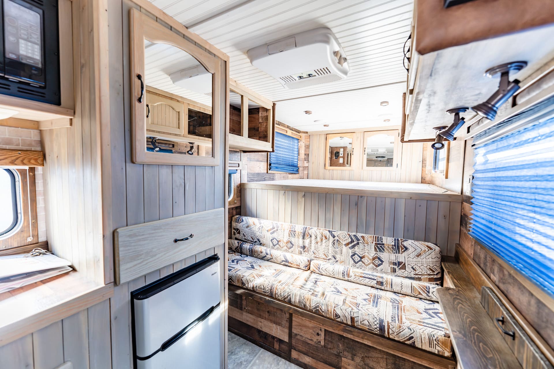 Wood-paneled RV living area with a patterned sofa, raised cabinets, and blue window blinds.