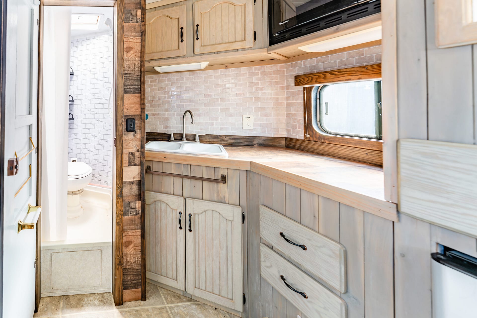 Compact kitchen in a rustic caravan or tiny home with light wood cabinets, tile backsplash, and a double sink under a window
