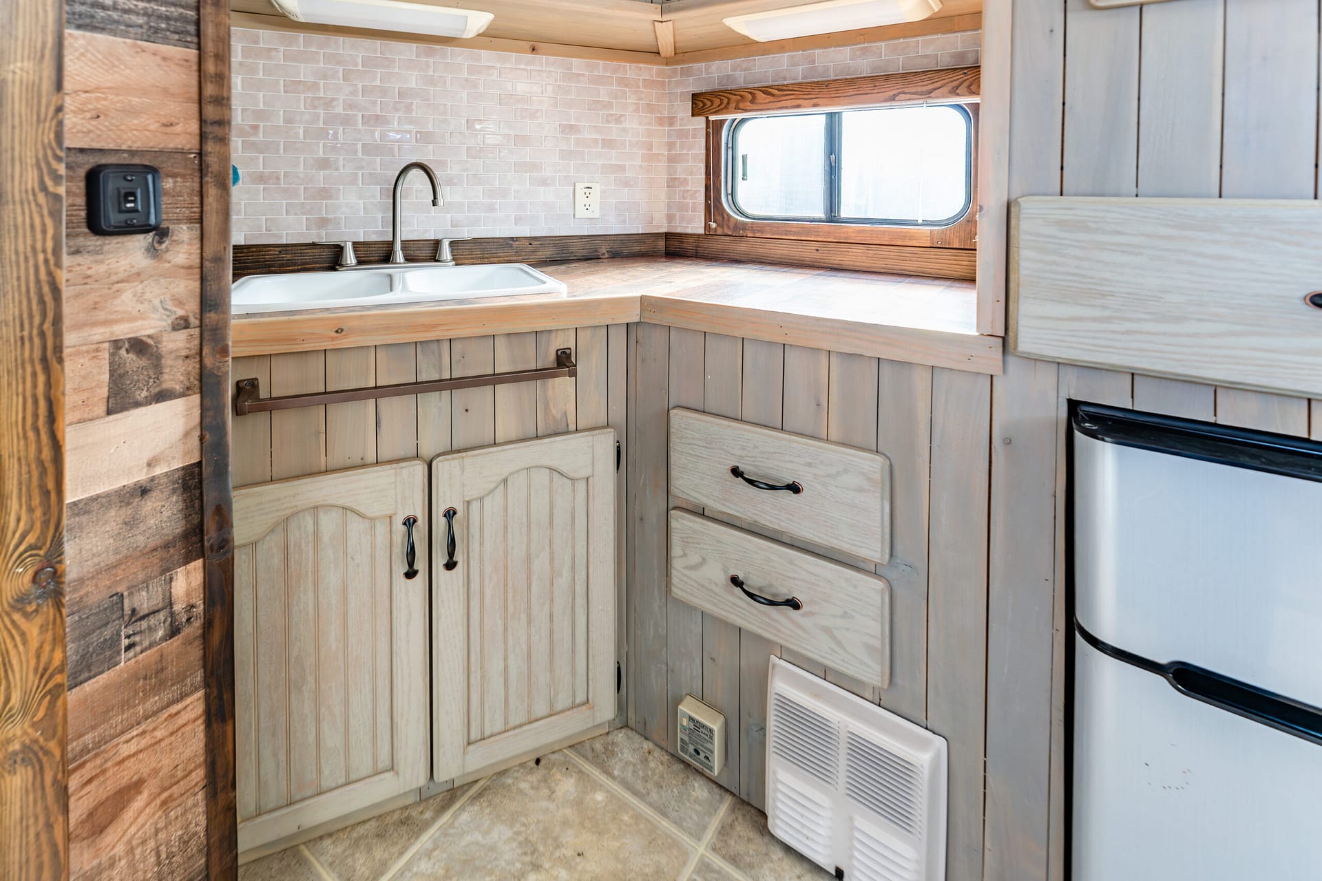 Rustic camper kitchen with a double sink, brushed-nickel faucet, and light wood cabinets under a beige tiled backsplash. A small window sits above the counter.