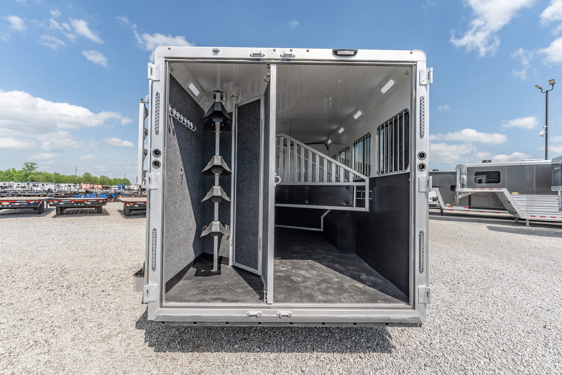 Open horse trailer rear with partitioned stalls and hooks, empty interior on a gravel lot under a blue sky