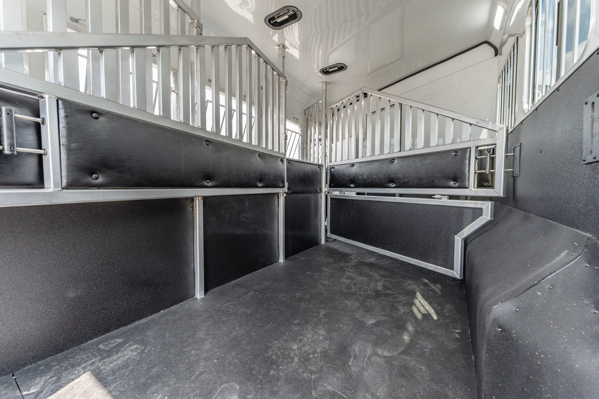 Interior of a metal horse trailer with partitioned stalls and black padded walls, gray aluminum rails, and a dark rubber floor in view