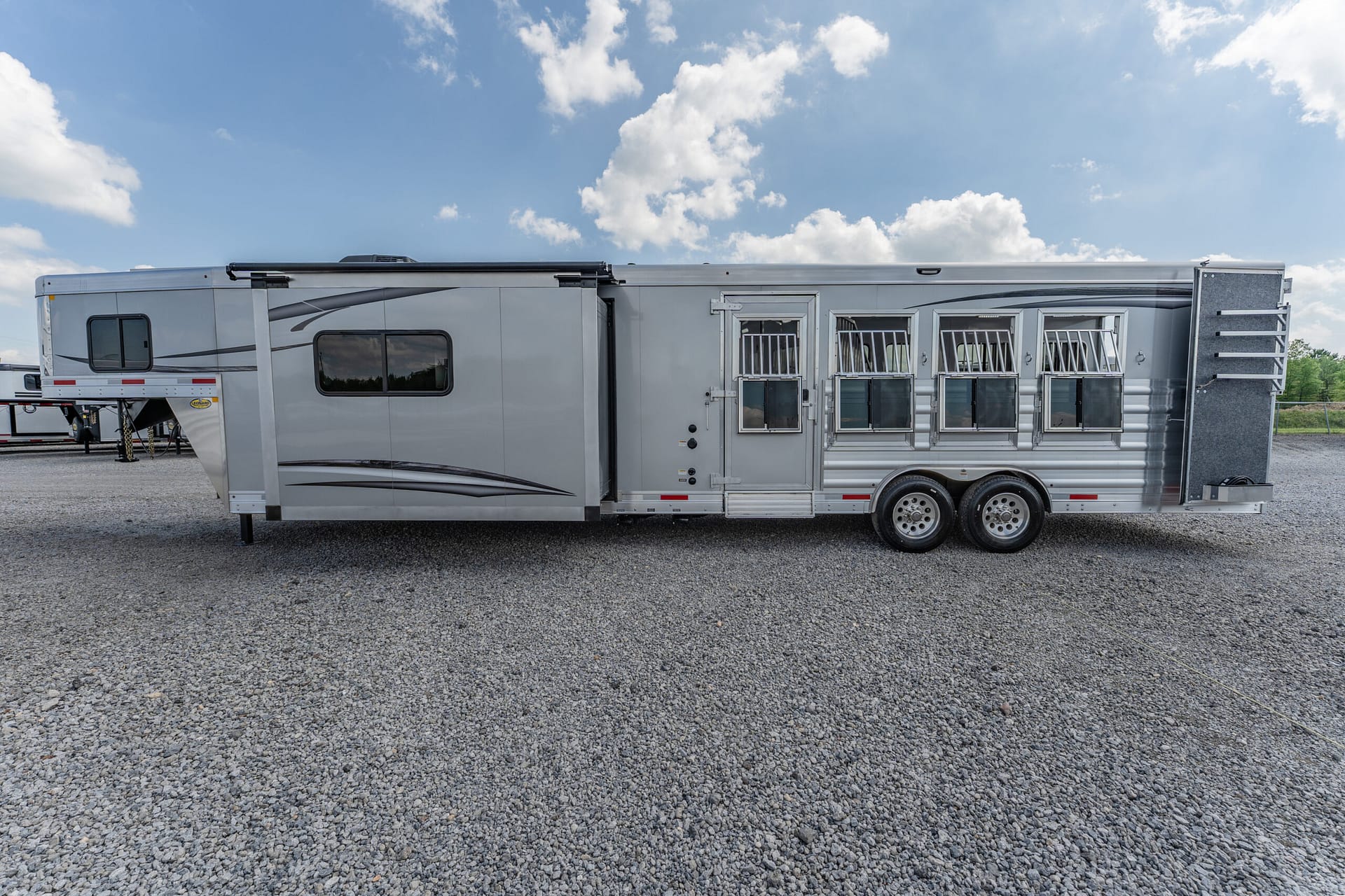 Side view of a long silver horse trailer with multiple stall windows parked on a gravel lot under a partly cloudy blue sky