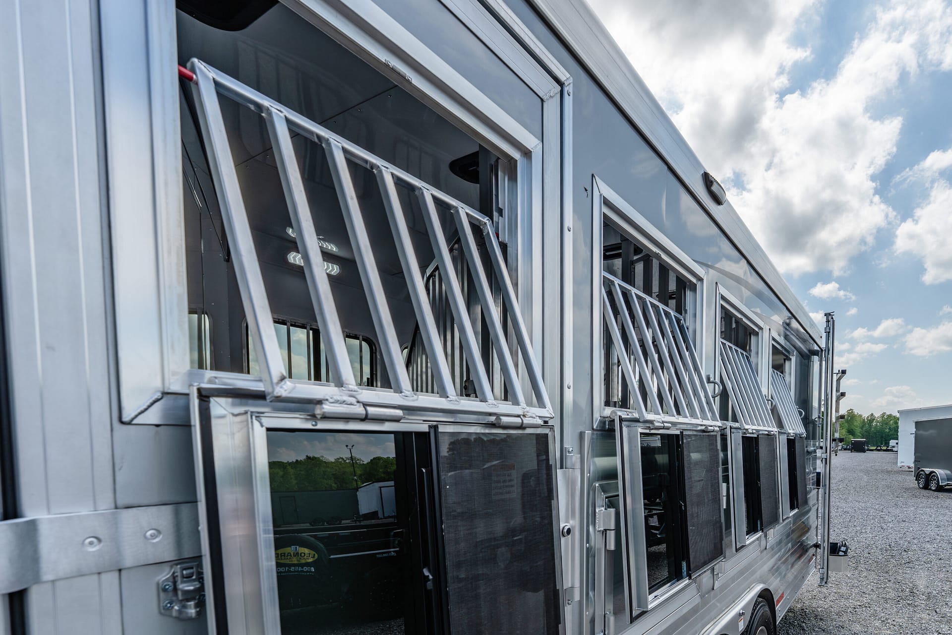 Silver tour bus exterior with attached metal ladders along the windows, parked on gravel under a cloudy sky.
