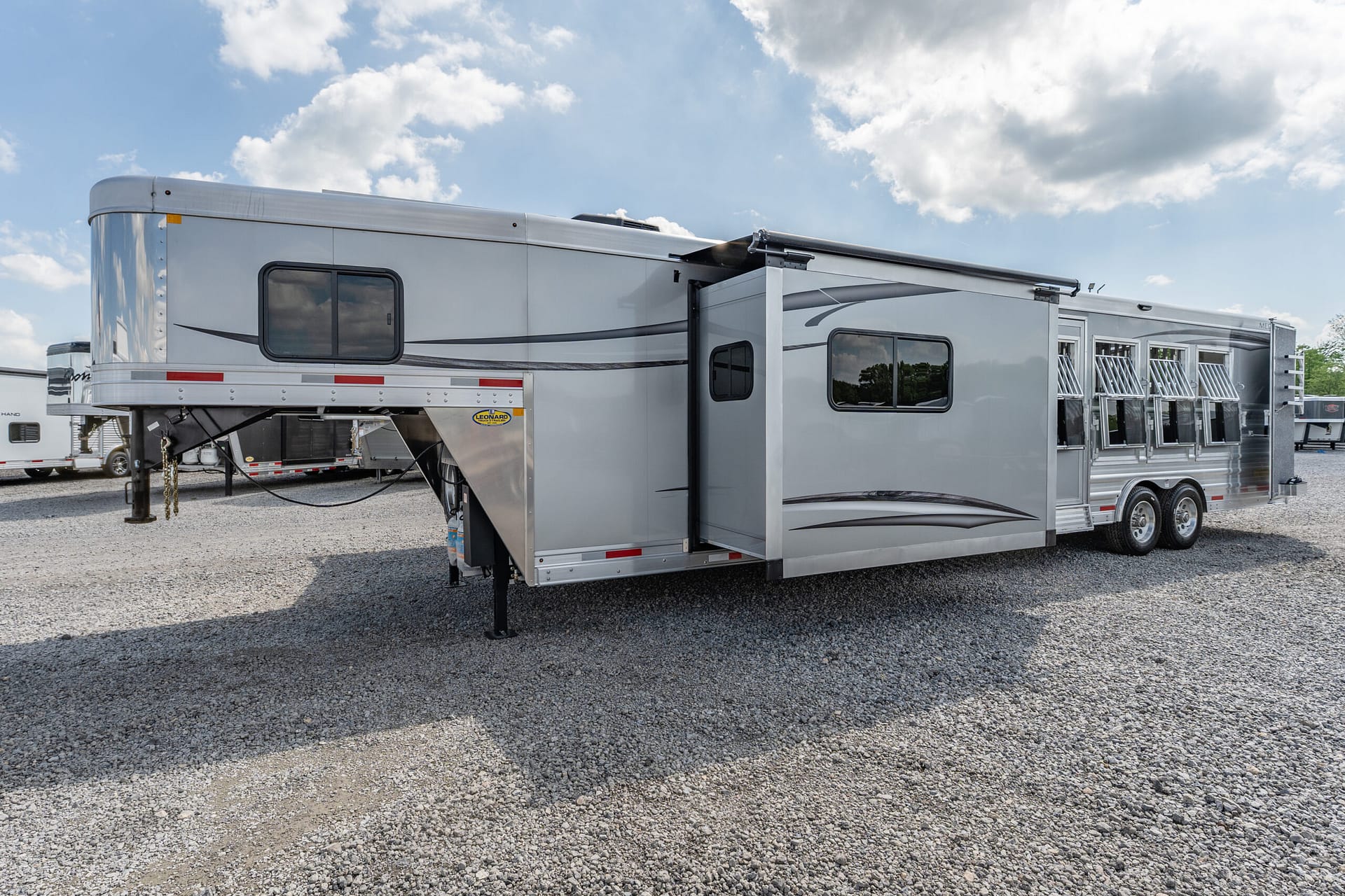 Silver fifth-wheel trailer on a gravel lot under a blue sky with clouds; slide-out section extended on the right side.