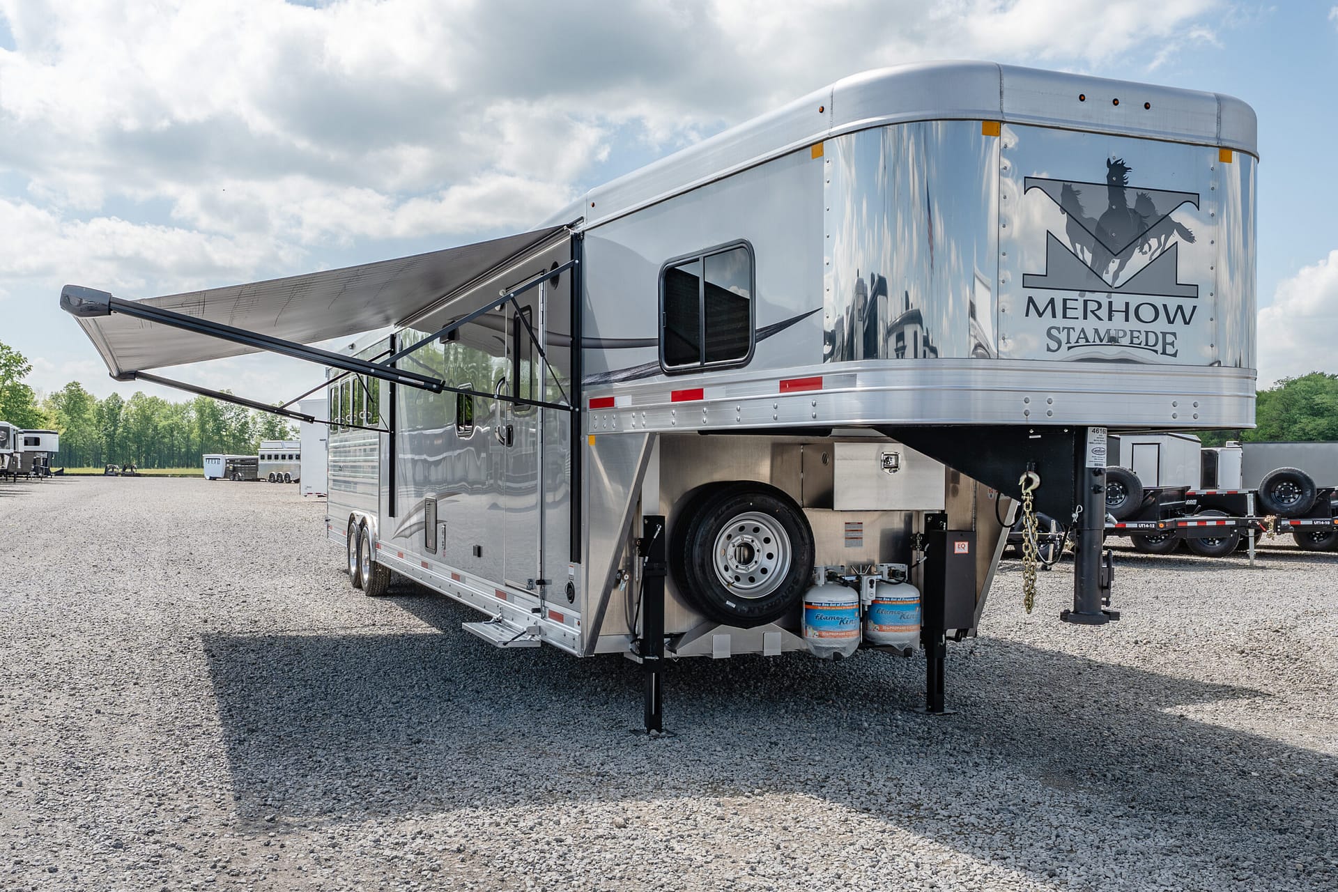 Shiny silver horse trailer with an extended black awning, parked on a gravel lot; Merhow Stampede logo on the rear door.