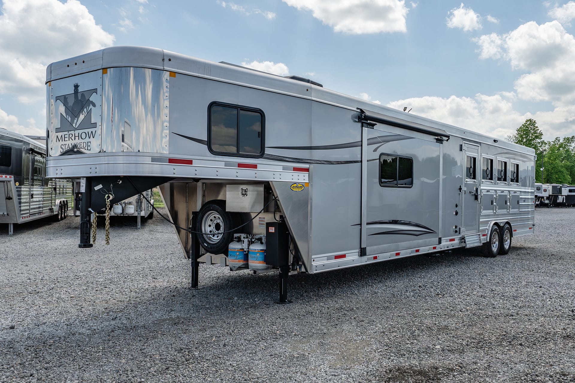 Silver horse trailer with a gooseneck hitch parked on a gravel lot; Merhow logo on the rear and a row of windows along the side, blue sky above.