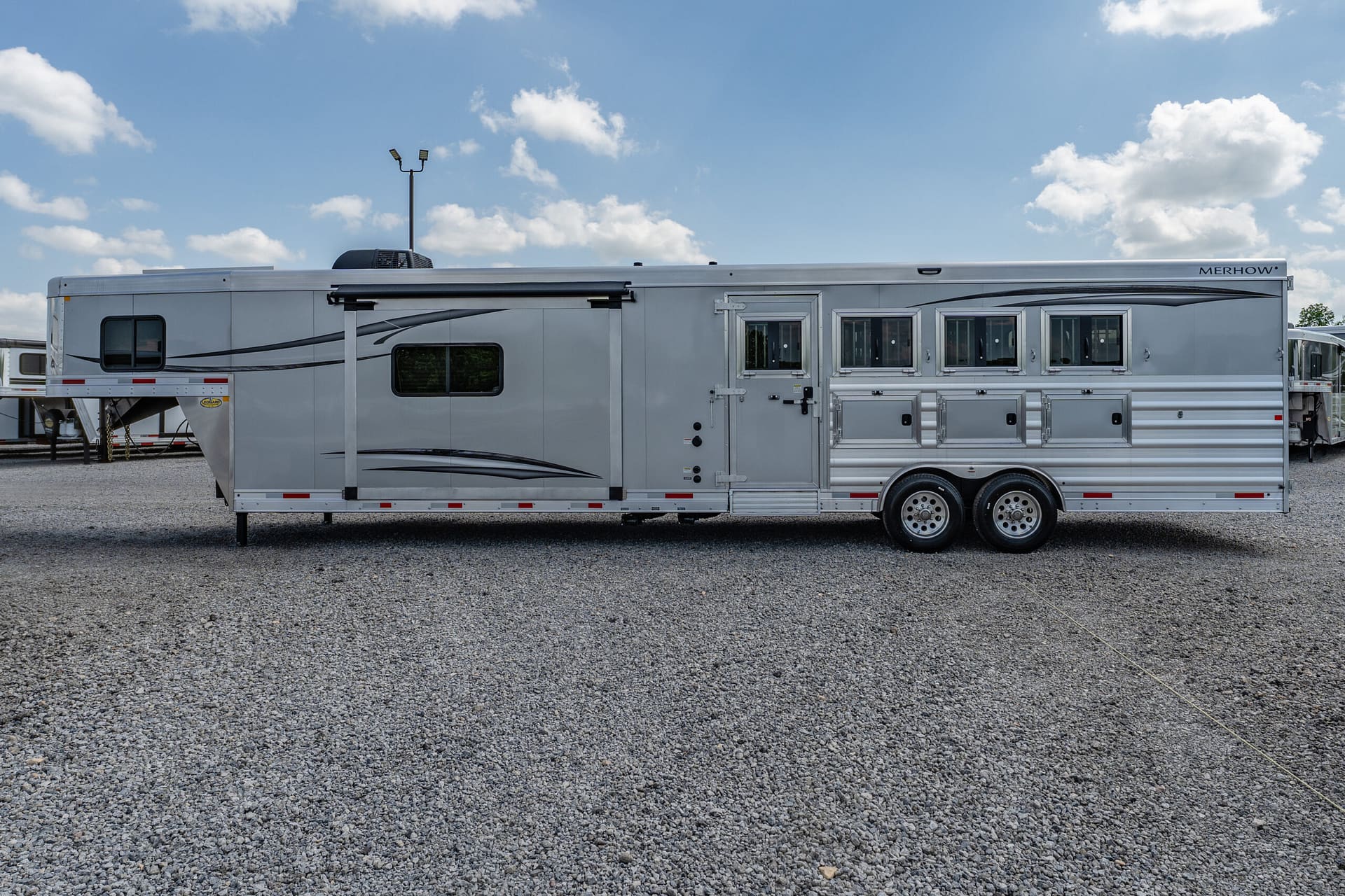 Long silver Merhow horse trailer parked on a gravel lot under a blue sky with scattered clouds.