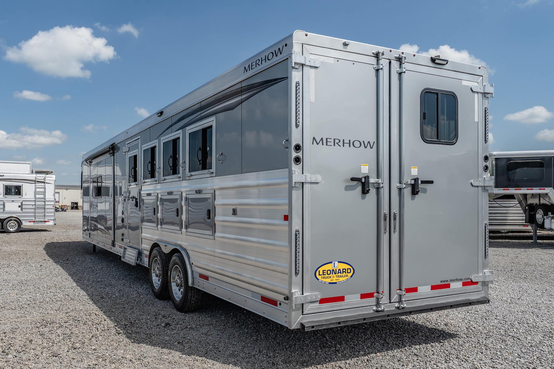 Large silver horse trailer with Merhow branding parked in a lot under a blue sky, Leonard trailer logo visible on the back.