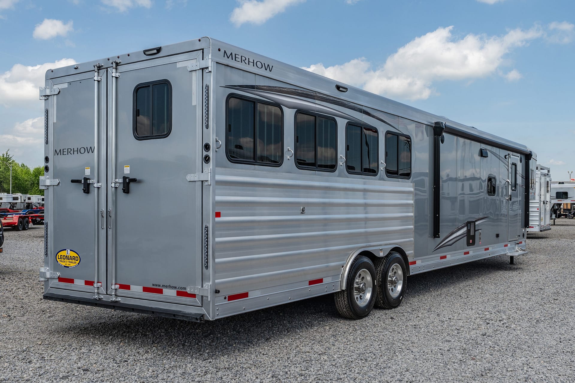 Rear-and-side view of a silver Merhow horse trailer with multiple windows, parked on gravel under a blue sky.