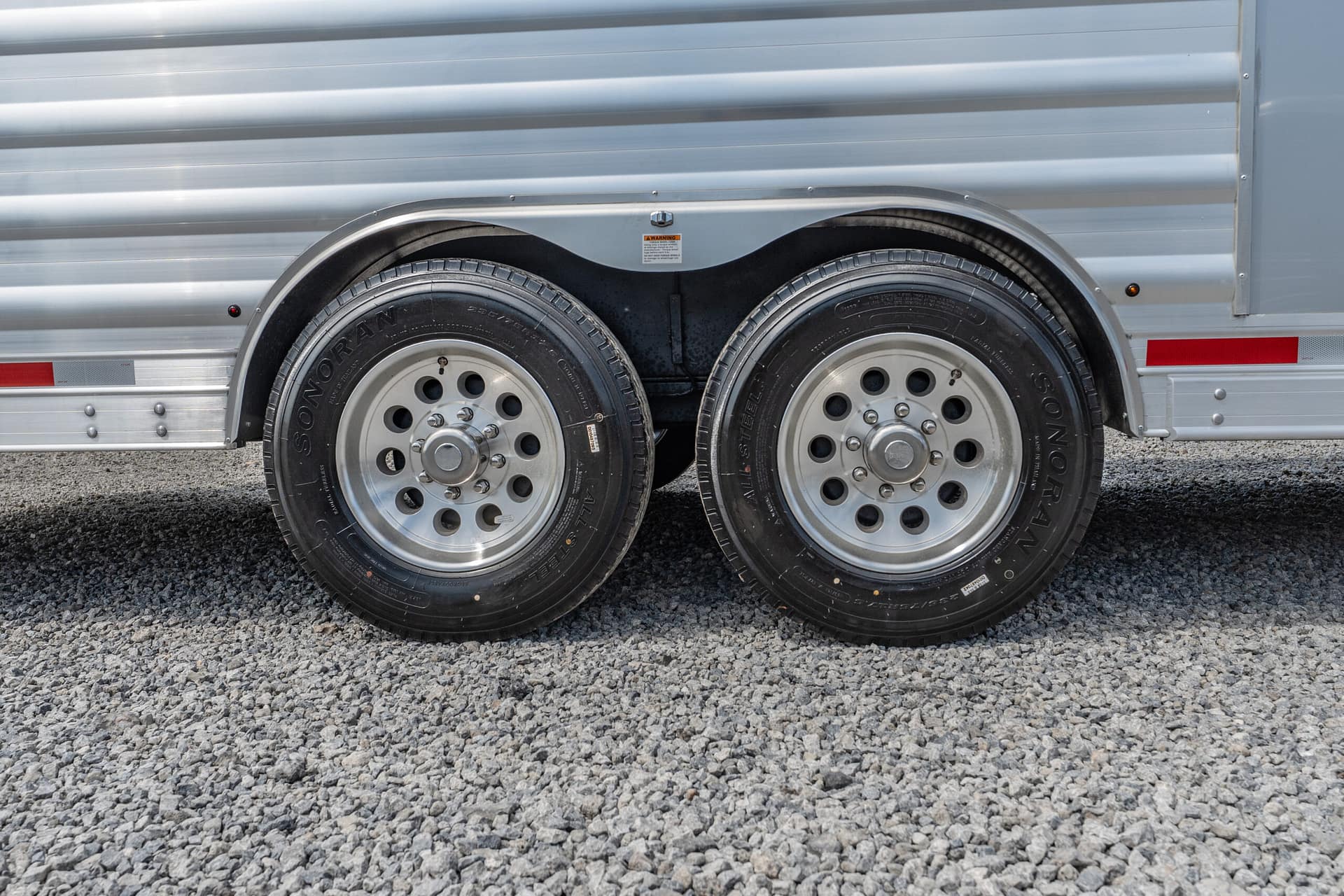 Close-up of two wheels on a silver aluminum trailer with a gravel surface beneath them, showing wheel wells and fenders.