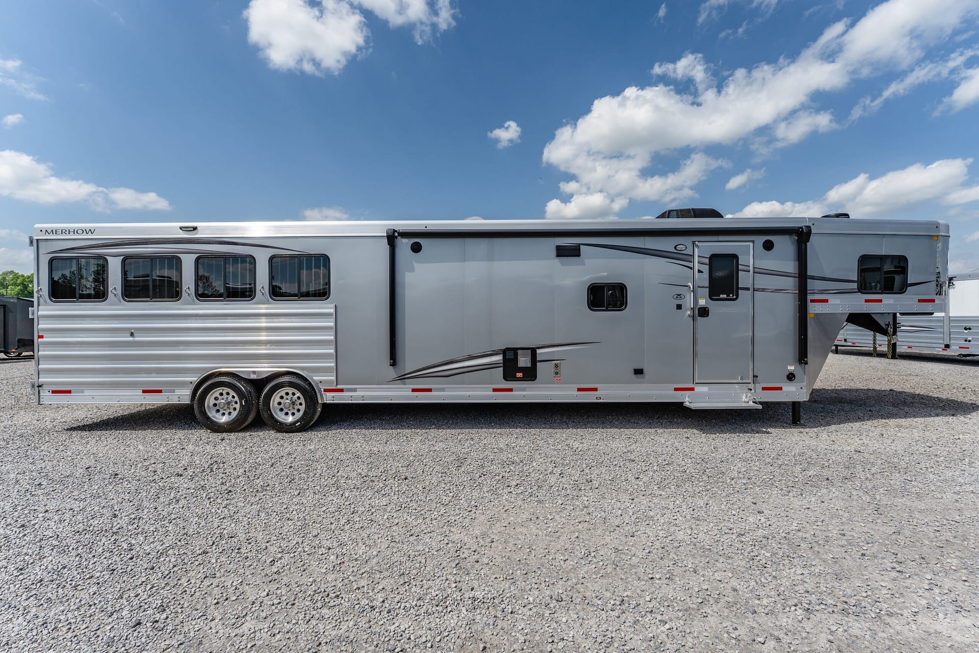 Silver Merhow horse trailer parked on a gravel lot under a blue sky with scattered clouds, side view with multiple windows and wheels.
