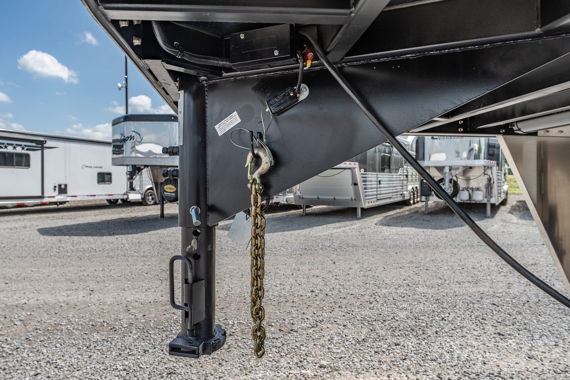 Close-up of a trailer tongue showing a hitch, safety chain, and wiring, with other trailers in a gravel lot behind it.