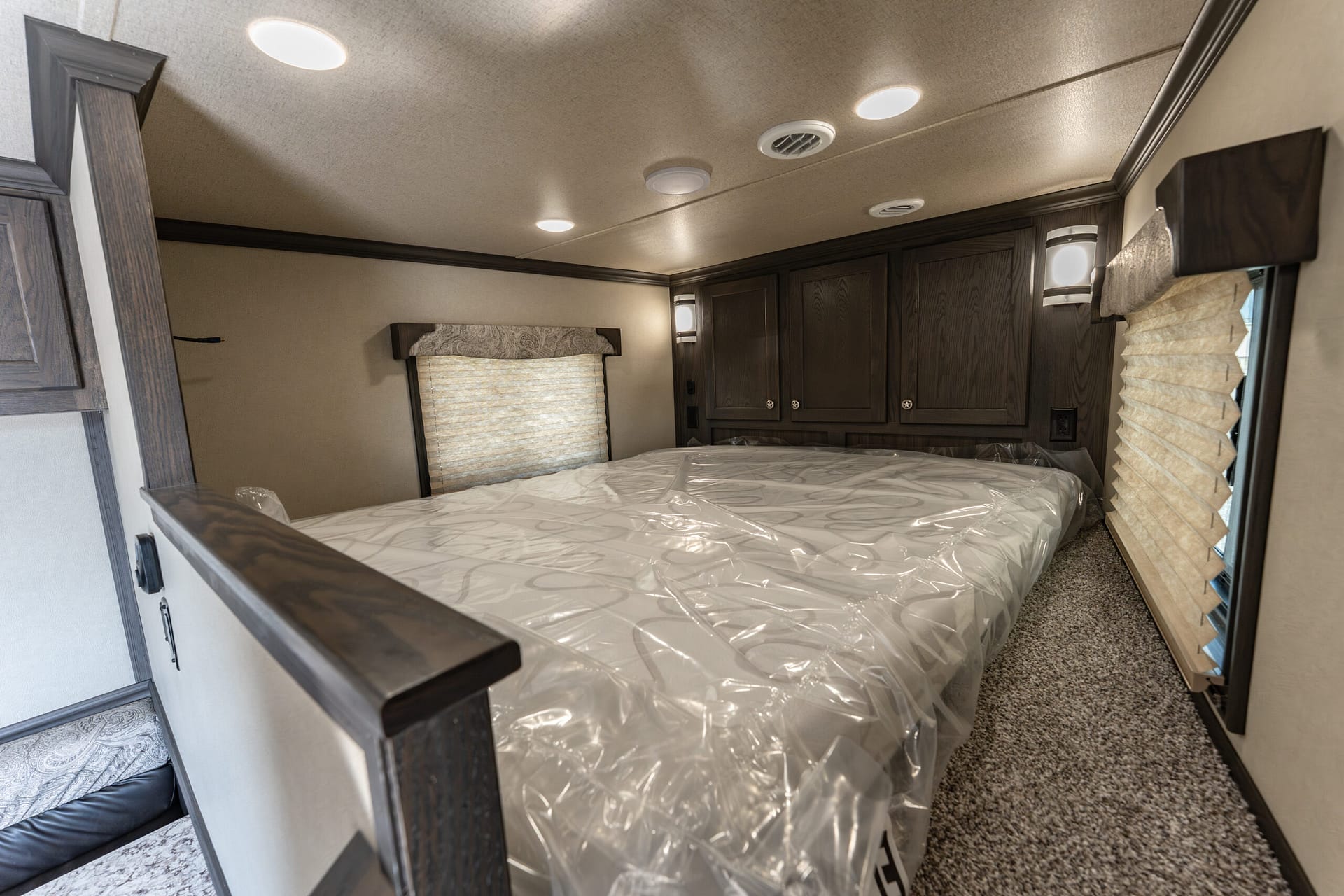 Compact RV bedroom with a full bed covered in plastic, dark wood cabinets above, and window blinds on either side.